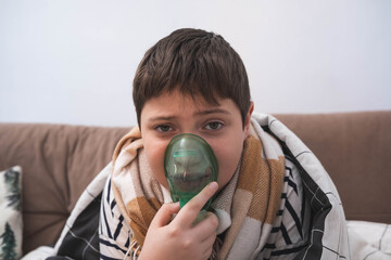 Boy using nebulizer mask while wrapped in blanket, sitting on sofa. Cozy indoor setting, focused expression, undergoing respiratory treatment at home.