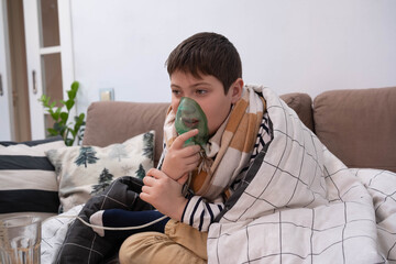 Boy wrapped in blanket using inhaler mask, sitting on sofa at home, recovering from illness, glass of water nearby, cozy indoor setting.