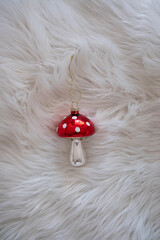 Red glass mushroom Christmas ornament with white dots on soft white fur background, festive holiday decoration, minimal winter flat lay.