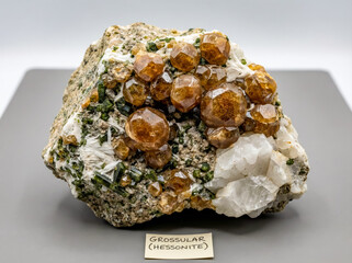 A collectible geological specimen of orange grossular garnet variety hessonite crystals on a skarn mineral matrix with green vesuvianite, fibrous wollastonite and large white calcite crystals.