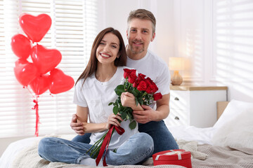 Happy couple with bouquet of roses on bed indoors. Valentine's day celebration