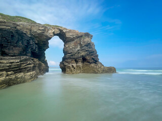 long exposure of a rock arch formation at Las Catedrales Beach in Ribadeo Galicia, Spain, with coastal cliffs, gentle waves, and a bright blue sky creating a stunning seascape