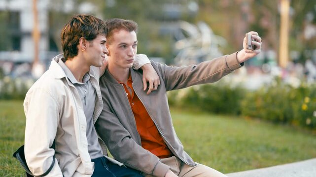 Two handsome young gay men sitting together in a park, taking a fun selfie with a smartphone. They are embracing, smiling, and making faces