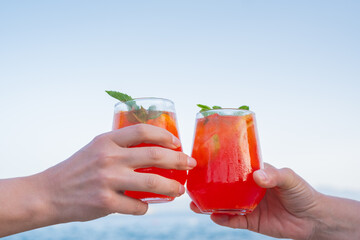 Two people raise glasses of red drinks by the sea at a resort during a sunny afternoon to celebrate their time together