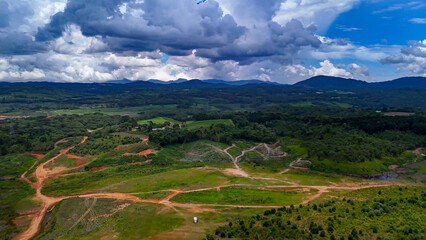 The image displays a vast rural and natural landscape with verdant hills, dense forest areas, cultivated fields, and a dramatic blue sky dotted with white and a few gray clouds.