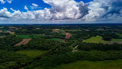 The image displays a vast rural and natural landscape with verdant hills, dense forest areas, cultivated fields, and a dramatic blue sky dotted with white and a few gray clouds.