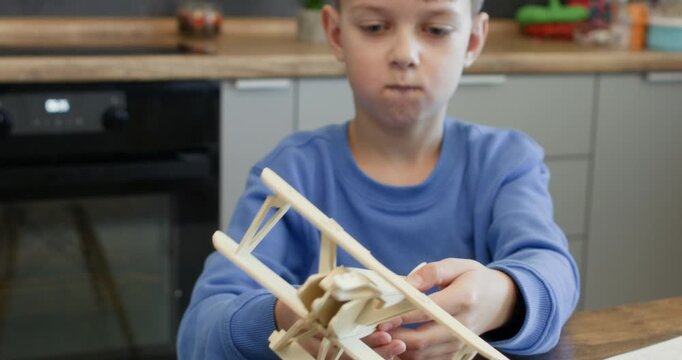Little boy shows unfinished wooden aeroplane model sitting at desk. Smart schoolboy plays with vintage toy biplane made of plywood construction at home