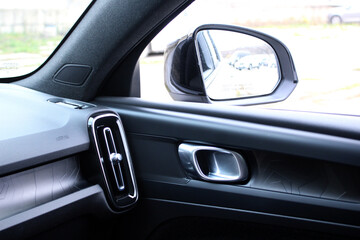 View of a modern car's interior from the passenger side, showcasing the black dashboard, vertical air vent, door handle, and a side-view mirror reflecting the exterior. Passenger airbag. Car air vent.