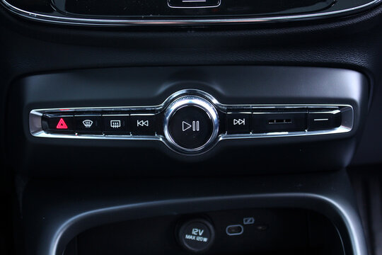 Close-up of a modern car's center console with media and function controls. The black panel features a central play and pause knob, hazard and defrost buttons, all highlighted by chrome accents.