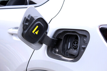Open electric car charging port on white vehicle body, plastic hinged lid with yellow warning symbol, black charging inlet with rubber seal, round connector socket, modern automotive design.