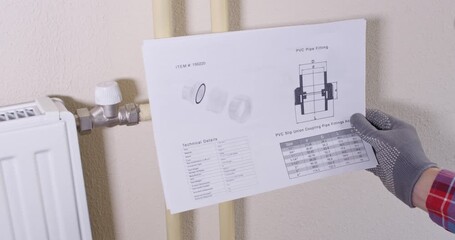 Gloved technician holds datasheet near light wall with radiator valves. Plumber positions ball valve according diagram while verifying orientation before installing