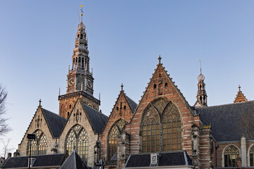 Low angle view of the Oude Kerk (Old Church) tower and gothic brick facade against a clear blue sky in the city center of Amsterdam Netherlands, 28 december 2025