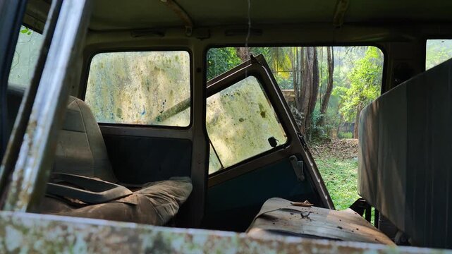 Interior of old abandoned van with broken seats and detached moss-covered door leaning inward showing decay and overgrown forest surroundings
