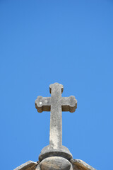 A weathered stone cross stands atop a traditional chapel roof against a vivid and cloudless blue sky background