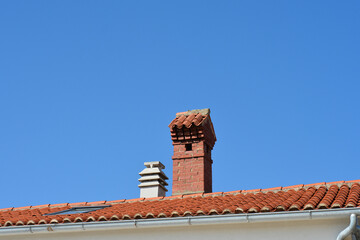 A tall decorative red brick chimney and a secondary white vent stand prominently on a Mediterranean house roof with classic orange clay tiles under a clear blue sky.