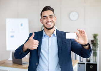 Happy Arab male entrepreneur holding empty business card, showing thumb up gesture at company office, mockup for design. Smiling CEO manager demonstrating blank advertising card
