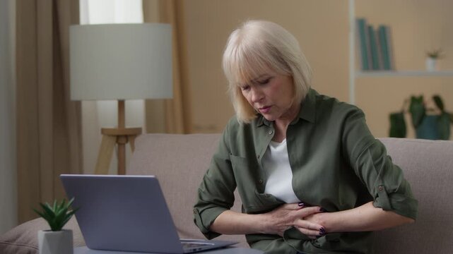 Old Caucasian woman with laptop at home feeling pain in stomach belly ache senior female retired lady sick ill bad feeling unhealthy granny with computer in living room bending holding abdominal spasm