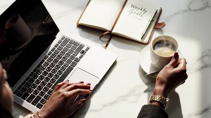 Overhead view of woman typing on laptop next to coffee and weekly goals notebook on marble desk