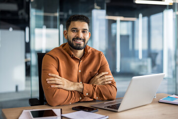 Young indian businessman sitting at desk in a modern office, looking at the camera with a confident, friendly smile, representing success, entrepreneurship, and professional corporate work