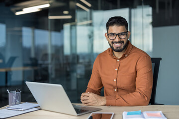 Young indian businessman with eyeglasses smiling at camera, sitting at desk with laptop in modern...