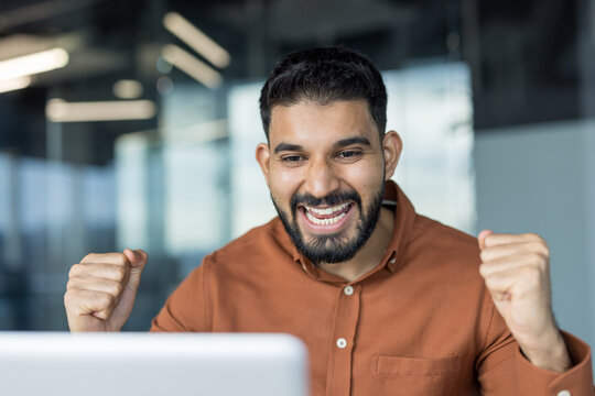Enthusiastic young man with a beard cheering and clenching fists while looking at a laptop screen, celebrating success or a win in a modern office environment