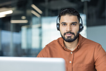 Indian man wearing a headset sitting at a desk, looking at the camera, working remotely or in a modern call center office providing client assistance and online technical support