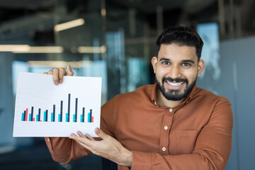 Young happy indian businessman smiling and holding a document with a bar chart, presenting business statistics, financial data, and positive growth in a modern office environment