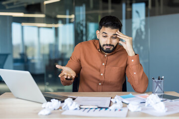 Young man experiencing stress and confusion while working on a laptop at a cluttered office desk, surrounded by crumpled paper, indicating struggles with assignments or creative block