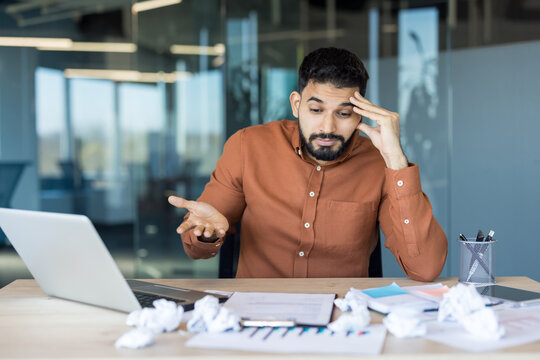 Young man experiencing stress and confusion while working on a laptop at a cluttered office desk, surrounded by crumpled paper, indicating struggles with assignments or creative block