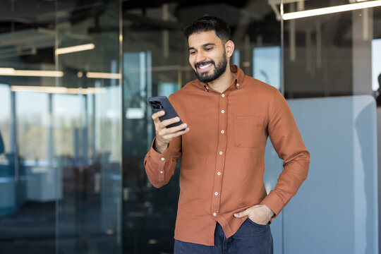 Indian man in a button-up shirt smiling and looking at his smartphone while standing in a modern office. Representing communication. Technology. And a connected lifestyle in a professional environment