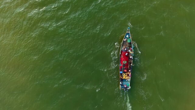 Fishing boat with red nets and crew navigating through deep green sea, creating foamy water trails captured from aerial view, symbolizing teamwork, labor, and traditional livelihood at sea
