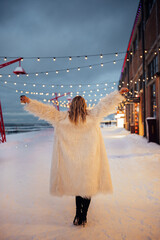 Woman walks on snowy street under string lights during winter evening by the coast
