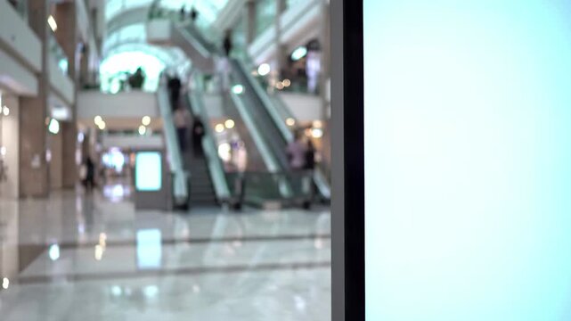 Blank digital advertising screen mockup in a modern shopping mall interior with blurred shoppers and escalators in the background