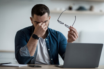 Tired young male feel pain eye strain hold glasses, rubbing dry irritated eyes, fatigued from computer work, stressed man suffer from headache, bad vision sight problem sit at home table using laptop