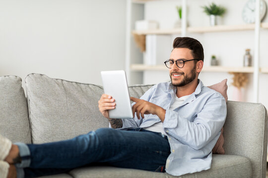 Technology concept. Smiling millennial man using digital tablet resting on couch at his modern apartment, free space. Happy guy browsing internet on pad, resting on sofa in living room
