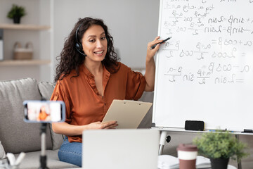 Studying Online During Covid-19 Pandemic. Smiling female math teacher in headphones giving lesson on web, using smartphone and pc. Millennial lecturer having video call with students recording vlog