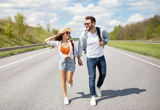Full length of positive young couple walking along highway, holding hands, having fun together outdoors. Cheerful millennial man with his girlfriend on road, hitchhiking, traveling by autostop