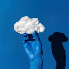 Holding a cloud made of cotton with a blue painted hand against a vibrant blue background near midday