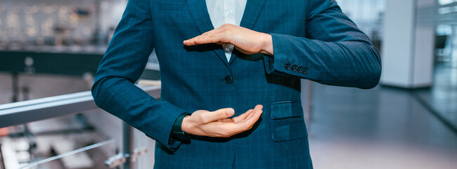 A person in a formal suit stands in a bright office. Their hands are positioned to show a...