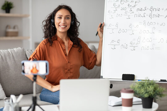 Online Learning Concept. Portrait of smiling female teacher in headset sitting near whiteboard, explaining math formulas to students. Excited woman posing and looking at camera, recording video class - Powered by Adobe