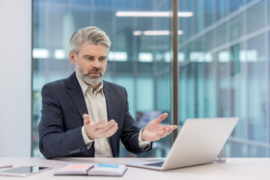 Mature businessman experiencing frustration and stress during a remote meeting or video conference, communicating difficulties while working on a laptop in a modern office setup