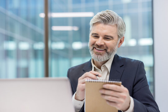 Mature gray-haired businessman close-up writing data in notebook, watching online video course webinar. Senior boss at online conference business meeting inside office. - Powered by Adobe