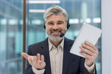 Mature call center agent looking at camera while working remotely from a modern office, wearing a headset and holding a tablet, providing online support and assistance