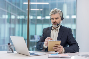 Middle-aged telemarketer wearing a headset and suit, concentrating while writing notes in a notebook during a remote online meeting in a modern office