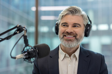 Mature man with a happy expression wearing headphones and speaking into a professional microphone,...