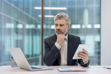 Experienced businessman evaluating corporate strategy, holding a digital tablet while sitting at a desk with a laptop, looking away in deep thought within a modern office setting