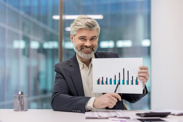 Smiling mature businessman holding up a bar chart, pointing to the data for analysis and illustrating positive financial growth and successful strategy in a modern office setting