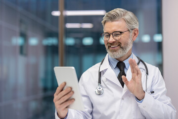 Mature doctor in white scrub uniform and stethoscope smiling, waving hand while having an online video call on a digital tablet, offering remote consultation and virtual care