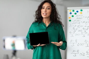 Modern Distance Learning. Female math teacher making video conference chat with students using cellphone on tripod, holding and showing tablet with empty screen with copy space for mock up, explaining