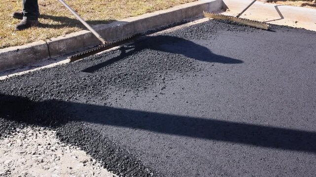 Workers use tools to spread hot asphalt on ground for road repair at construction site
